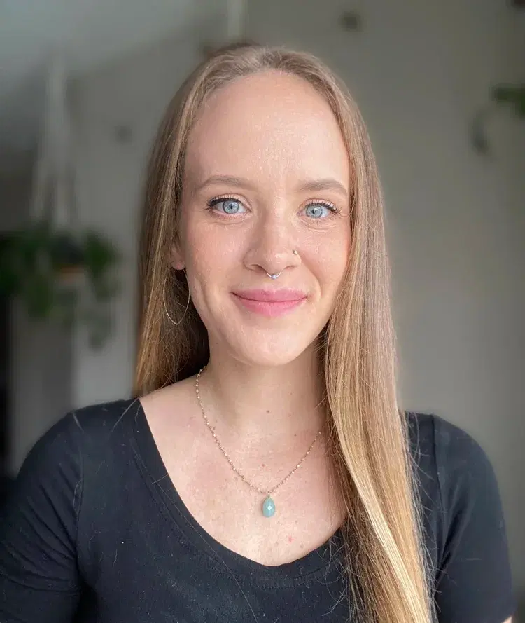 Close-up portrait of a woman with long light brown hair, blue eyes, and fair skin, smiling at the camera in a well-lit room, wearing a black top, a gold necklace with a blue pendant, and a nose piercing.